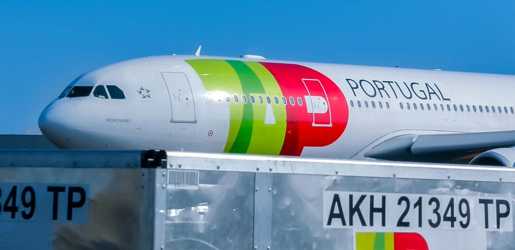 A imagem mostra a parte frontal de um avião Airbus A330-200 da TAP Air Portugal. O avião exibe a pintura característica da TAP, com faixas em verde e vermelho formando o logotipo da empresa junto à palavra "PORTUGAL" escrita em letras maiúsculas. Na fuselagem, próximo ao cockpit, está escrito "PEDRO NUNES", o nome atribuído ao avião. Em primeiro plano, há um contentor metálico de carga identificado com o código "AKH 21349 TP", que também exibe o logotipo colorido da TAP. O céu ao fundo está limpo. A imagem mostra a parte frontal de um avião Airbus A330-200 da TAP Air Portugal. O avião exibe a pintura característica da TAP, com faixas em verde e vermelho formando o logotipo da empresa junto à palavra "PORTUGAL" escrita em letras maiúsculas. Na fuselagem, próximo ao cockpit, está escrito "PEDRO NUNES", o nome atribuído ao avião. Em primeiro plano, há um contentor metálico de carga identificado com o código "AKH 21349 TP", que também exibe o logotipo colorido da TAP. O céu ao fundo está limpo.