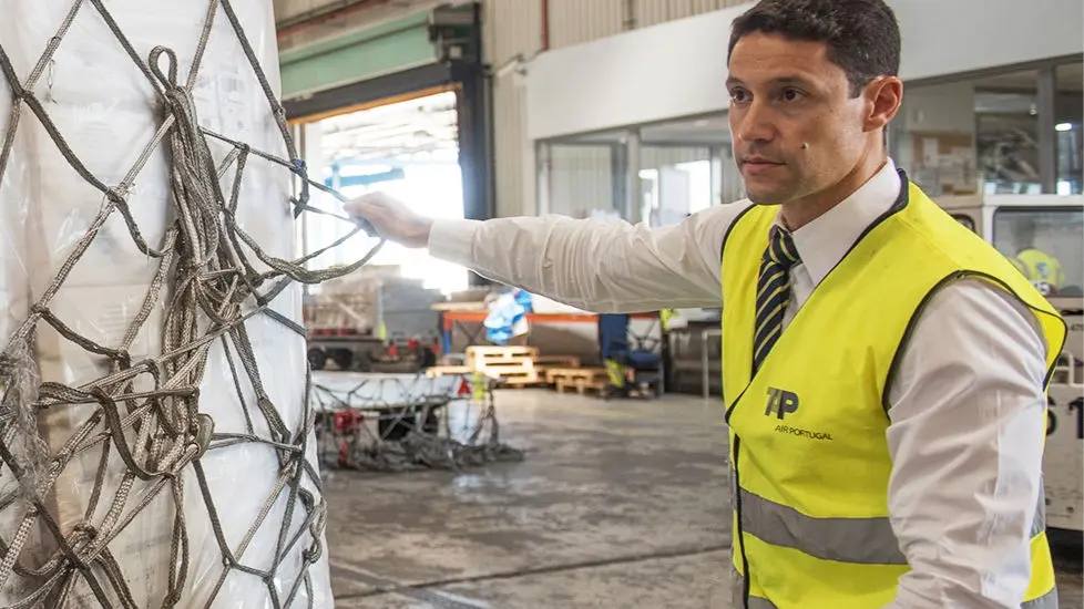 Um trabalhador da TAP Air Cargo com um colete refletor amarelo a manusear uma palete de carga aérea envolta por uma rede de contenção, num terminal aeroportuário.