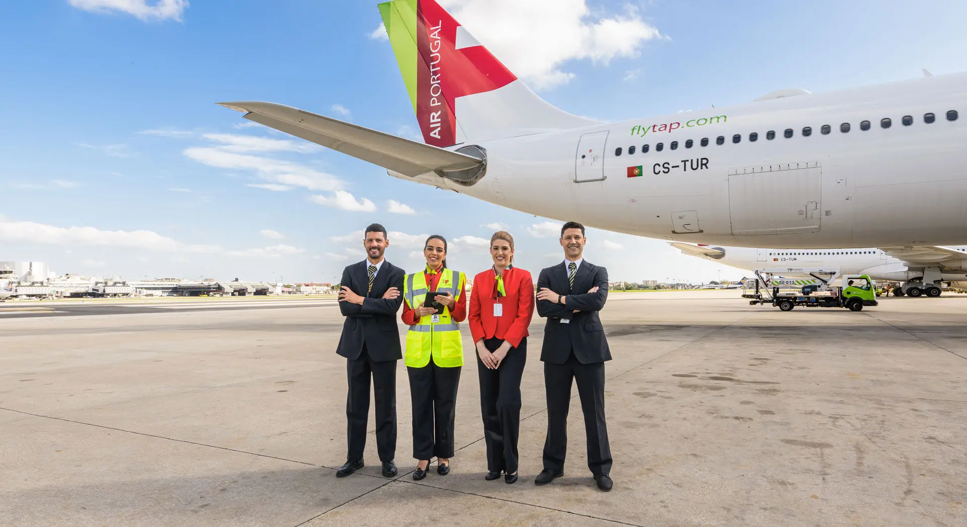 Quatro elementos (dois homens nas pontas e duas mulheres ao centro) da equipa de carga da TAP Air Portugal, em pé, na pista de um aeroporto, junto a uma aeronave da empresa. A cauda do avião exibe o logotipo e as cores da TAP Air Portugal (vermelho e verde), bem como a inscrição “flytap.com” e a matrícula da aeronave CS-TUR, que corresponde a um Airbus A330-900neo.