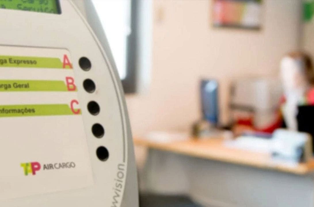 Ambiente de atendimento ao Cliente da TAP Air Cargo. Em primeiro plano é visível um painel de senhas eletrónicas com o logo da TAP Air Cargo. O painel tem letras que indicam diferentes serviços: A - Carga Expresso; B - Carga Geral; C - Informações. Em segundo plano, desfocado, é visível uma funcionária da TAP sentada numa secretária com um blazer vermelho e lenço verde-lima ao pescoço.