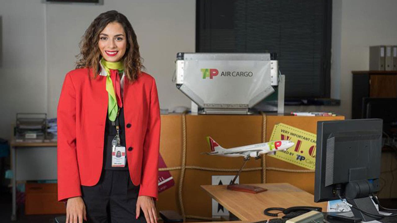 Uma funcionária do setor TAP Air Cargo, da TAP Air Portugal, pousa para uma fotografia num ambiente de escritório. A mulher tem os cabelos ondulados e veste um uniforme corporativo da TAP Air Portugal composto por um blazer vermelho , um lenço verde-lima atado ao pescoço, e umas calças de fato pretas. Ao fundo é visível uma unidade de carga com a marca TAP Air Cargo  sobre uma caixa simulada de carga aérea com uma etiqueta de "Very Important Cargo. É ainda visível uma maqueta de um avião da TAP sobre uma secretária de trabalho.