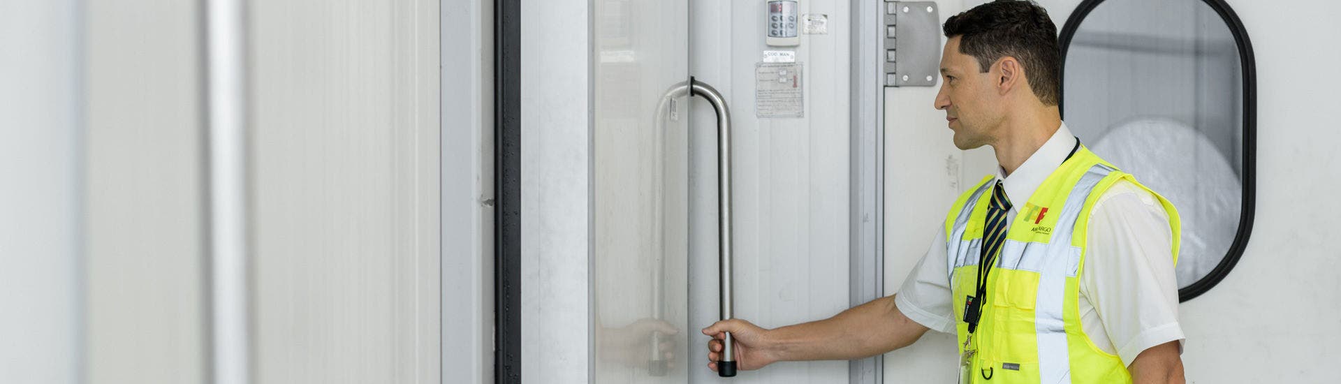 Funcionário com colete refletor amarelo e camisa branca abre uma porta metálica numa área industrial ou de armazém, com visor redondo visível ao fundo.