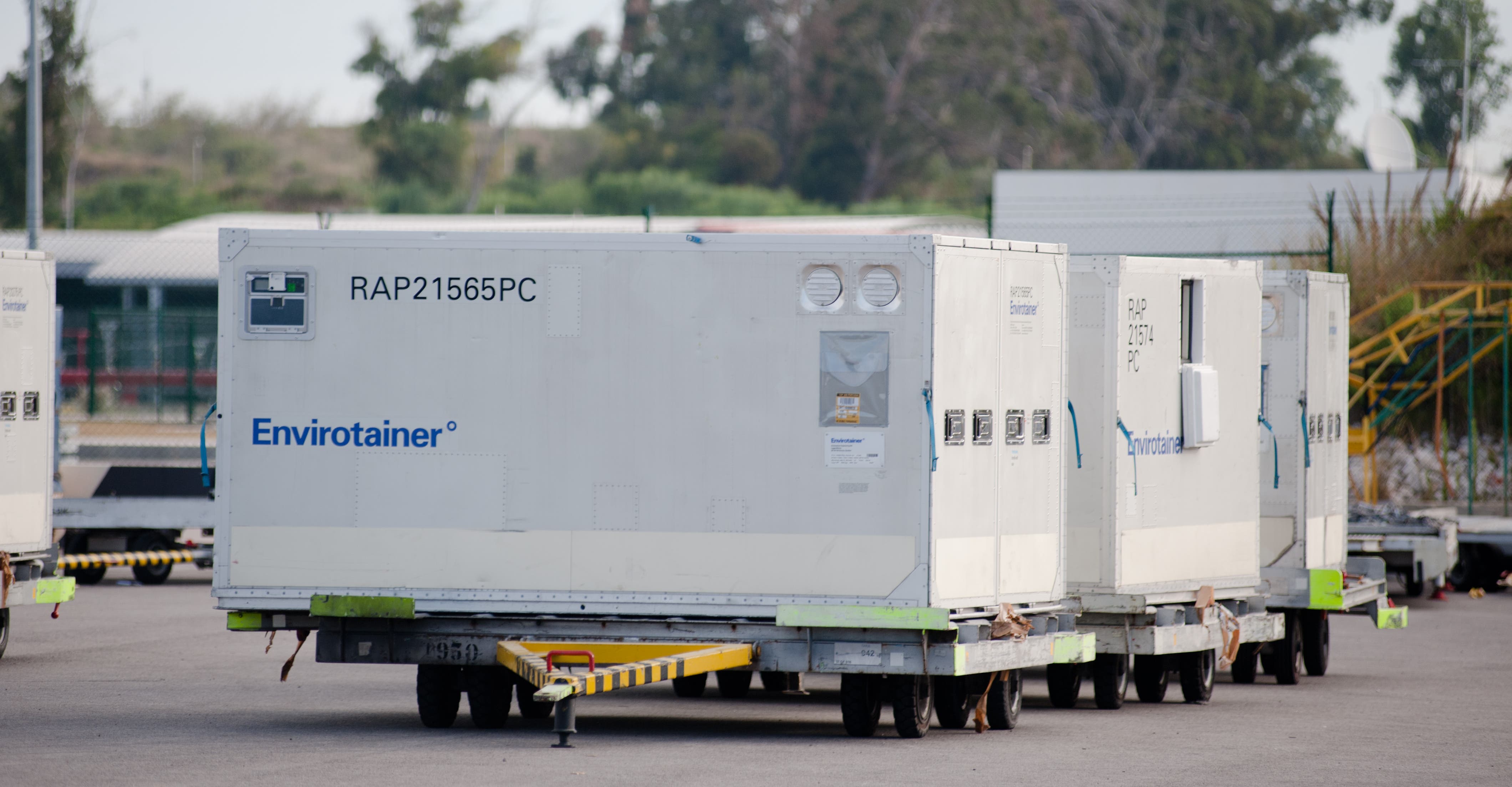 Contentores de transporte refrigerados, conhecidos como Envirotainer, alinhados sobre plataformas com rodas numa área de movimentação de carga, num aeroporto. Os contentores são de grande porte, de cor branca, e têm inscrições azuis com a marca Envirotainer e códigos de identificação como RAP21565PC e RAP21574PC. Estes contentores são utilizados para o transporte de produtos sensíveis à temperatura, como medicamentos, vacinas ou produtos farmacêuticos. Ao fundo, vê-se uma vedação e vegetação.