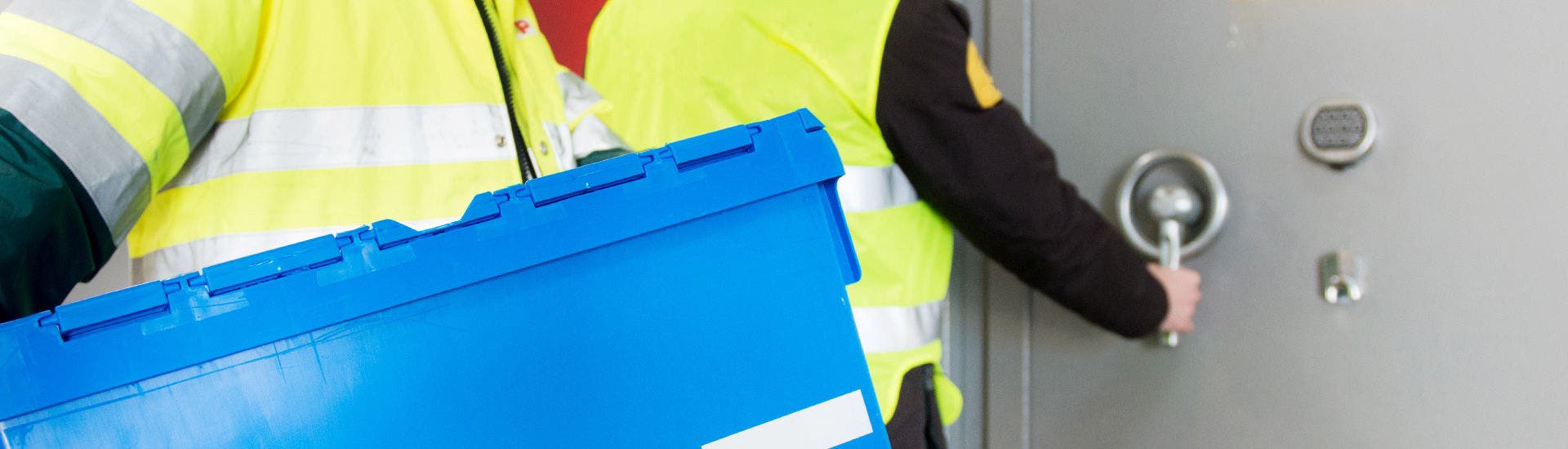 Um trabalhador da TAP Air Cargo transporta uma caixa plástica azul, normalmente usada para transporte de documentos ou bens. Atrás um segurança abre uma porta blindada de um cofre.
