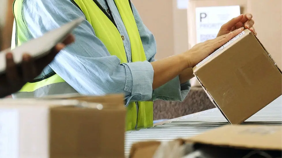 Um funcionário da TAP Air Cargo com um colete refletor amerelo manuseia uma caixa de cartão sobre um tapete de roldanas. Em primeiro plano, vêem-se outras caixas de cartão e, no canto inferior esquerdo, outra pessoa segura um caderno.