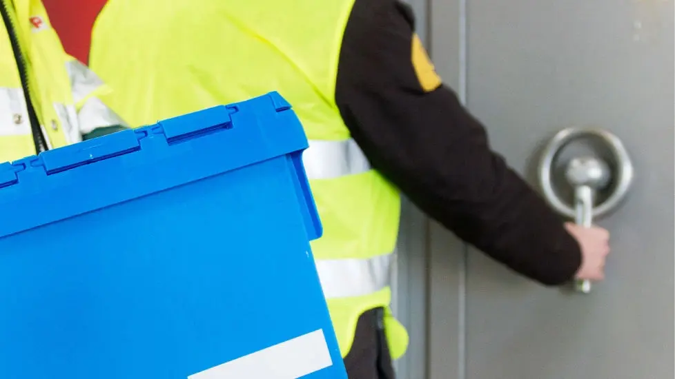 Um trabalhador da TAP Air Cargo transporta uma caixa plástica azul, normalmente usada para transporte de documentos ou bens. Atrás um segurança abre uma porta blindada de um cofre.