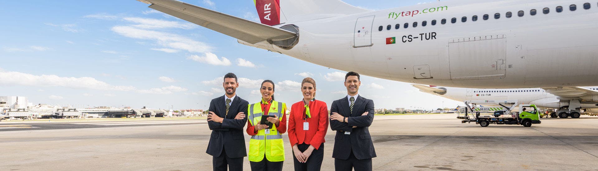 Quatro funcionários da TAP Air Portugal, com uniformes formais e colete refletor, posam juntos em frente a um avião da TAP Air Portugal estacionado na placa do aeroporto durante o dia, com céu azul ao fundo.