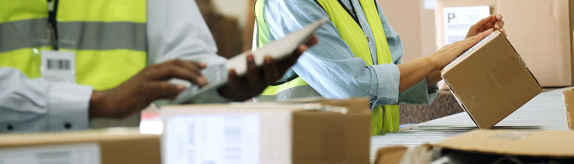 Um funcionário da TAP Air Cargo com um colete refletor amerelo manuseia uma caixa de cartão sobre um tapete de roldanas. Em primeiro plano, vêem-se outras caixas de cartão e, no canto inferior esquerdo, outra pessoa segura um caderno.