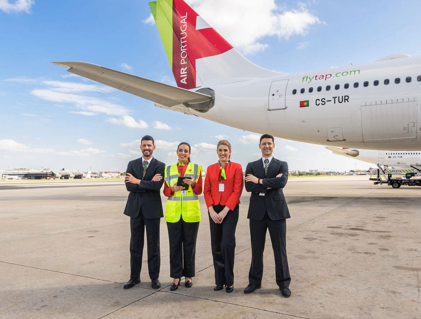 Quatro elementos (dois homens nas pontas e duas mulheres ao centro) da equipa de carga da TAP Air Portugal, em pé, na pista de um aeroporto, junto a uma aeronave da empresa. A cauda do avião exibe o logotipo e as cores da TAP Air Portugal (vermelho e verde), bem como a inscrição “flytap.com” e a matrícula da aeronave CS-TUR, que corresponde a um Airbus A330-900neo.