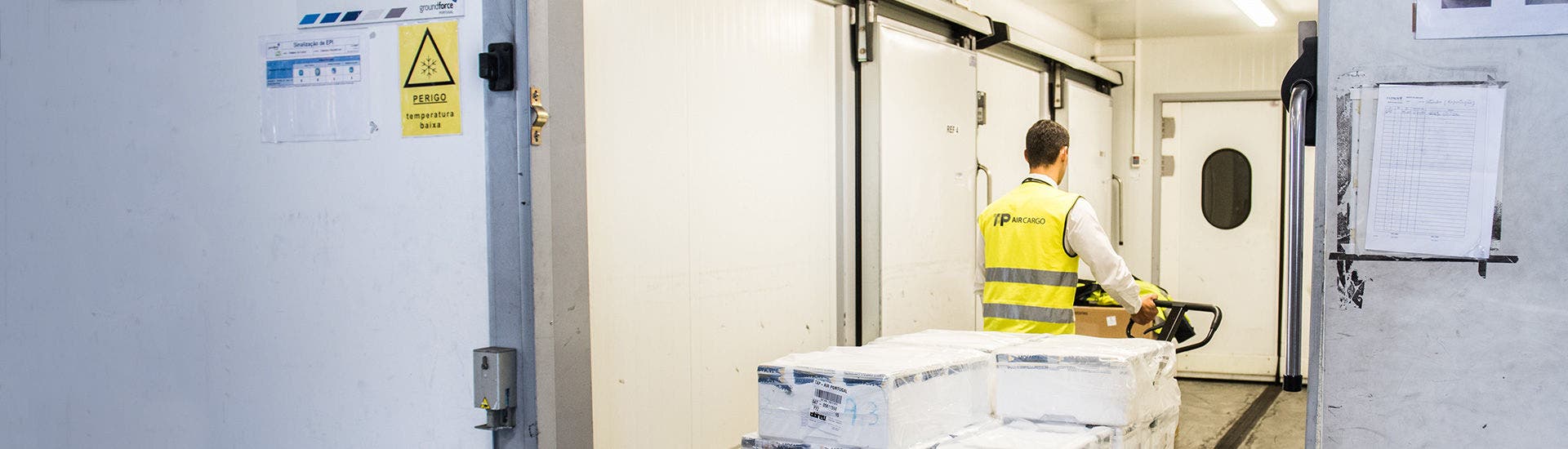 A TAP Air Cargo employee wearing a reflective vest maneuvers, with his back turned, a hand pallet truck carrying several cargo packages wrapped in plastic for protection.  This is done at a cargo storage area at an airport.