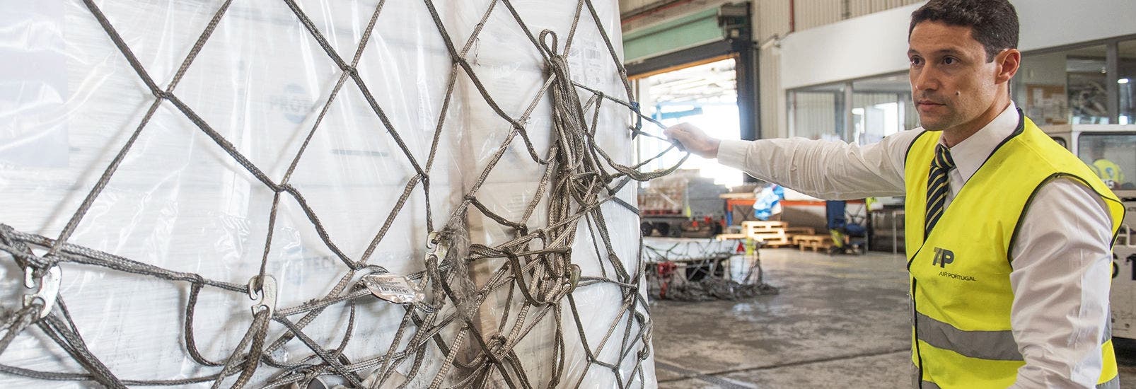 Um trabalhador da TAP Air Cargo com um colete refletor amarelo a manusear uma palete de carga aérea envolta por uma rede de contenção, num terminal aeroportuário.
